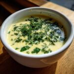 Zbliżenie na kremową zupę kalafiorową z koperkiem, podaną w białej miseczce na drewnianym stole.