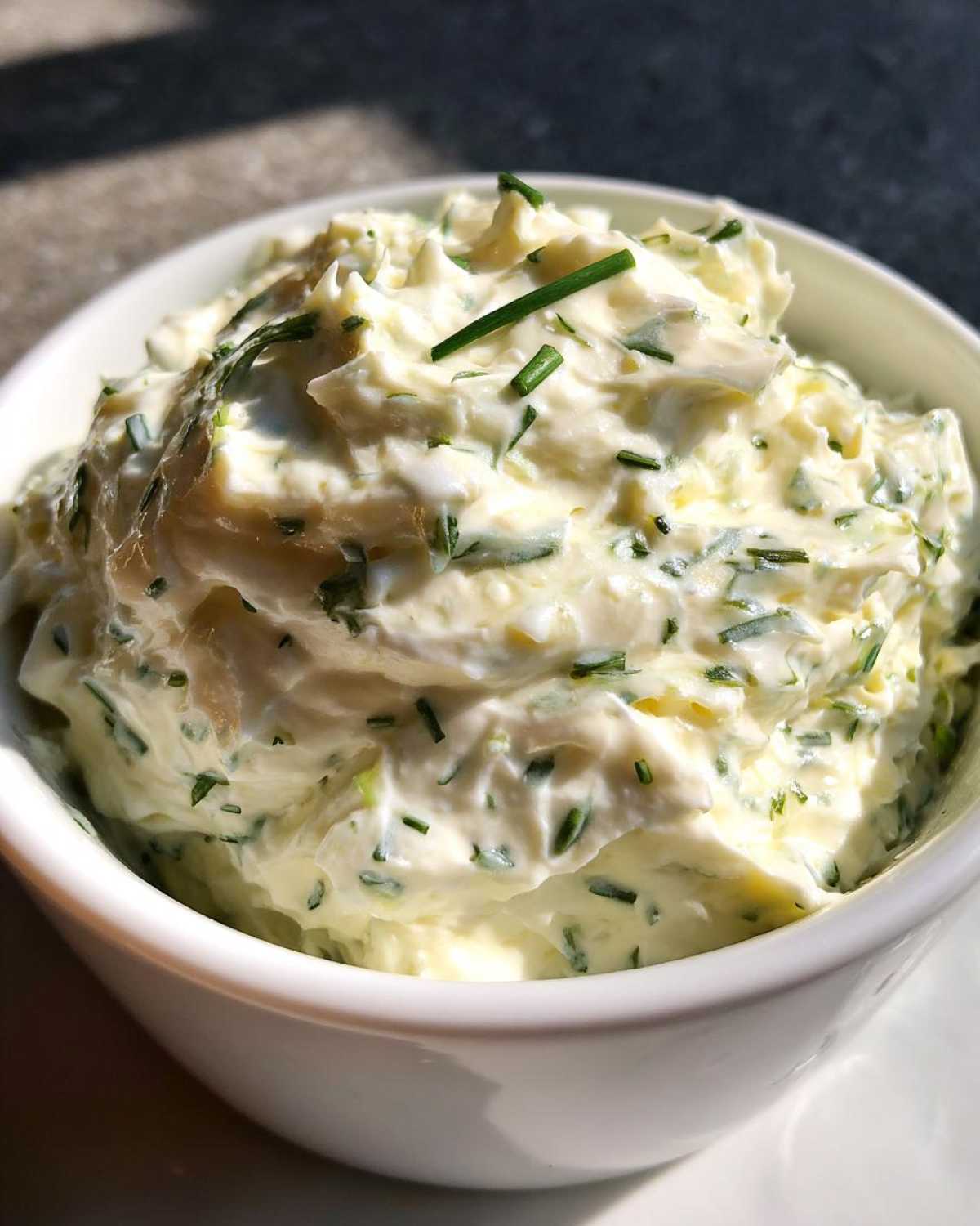 Zbliżenie na białą miseczkę wypełnioną kremowym dipem czosnkowym z widocznymi kawałkami zielonego szczypiorku.