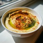 Gładki, kremowy Hummus klasyczny w białej misce, polany oliwą, papryką i posypką.