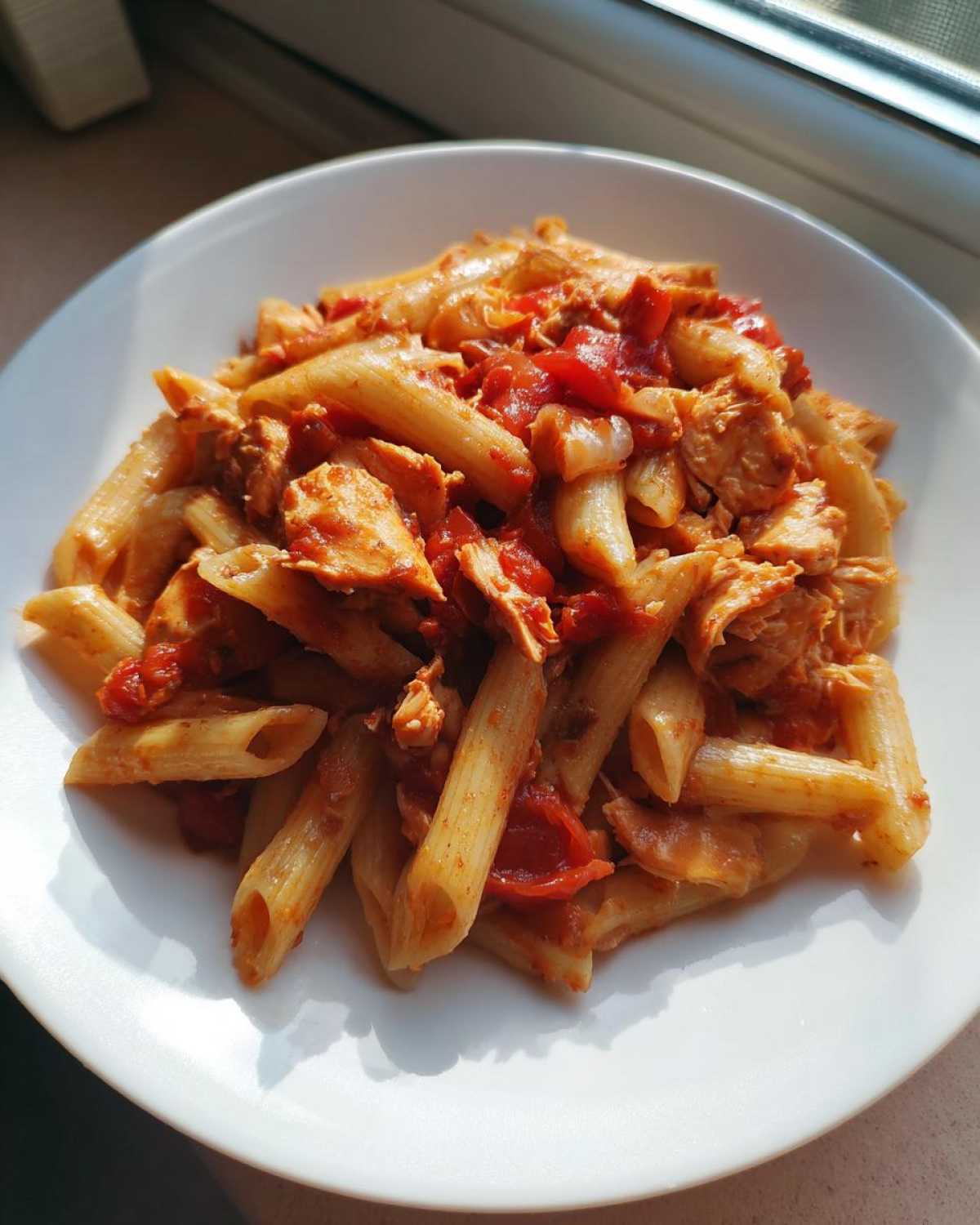 Zbliżenie na biały talerz z porcją makaronu penne z sosem pomidorowym i tuńczykiem, oświetlone słońcem.