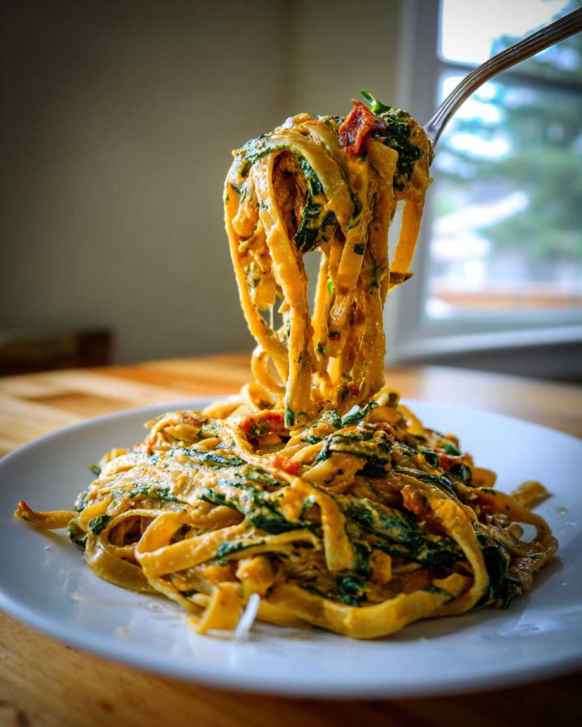 Kluski makaronu typu tagliatelle z sosem, szpinakiem i suszonymi pomidorami, podnoszone widelcem z białego talerza. Idealny makaron ze szpinakiem i suszonymi pomidorami.