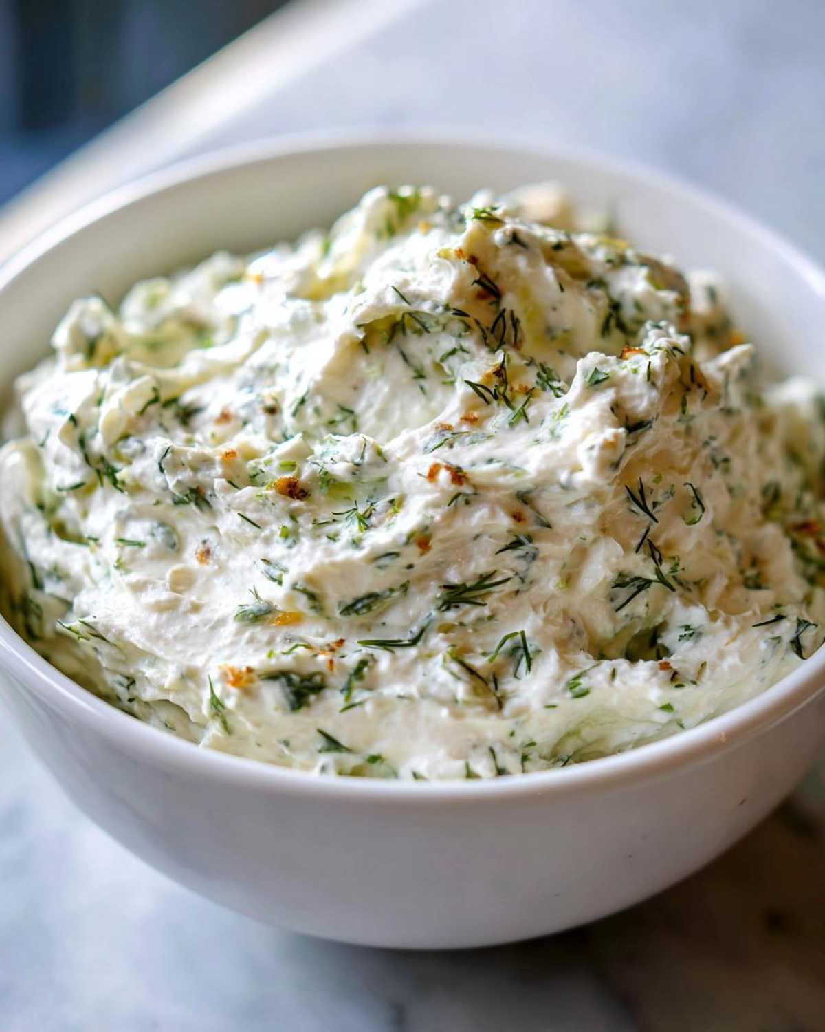 Zbliżenie na kremowy, puszysty dip czosnkowy z dużą ilością posiekanego koperku w białej misce.