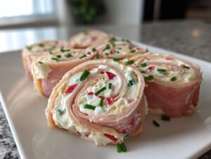 Zbliżenie na Roladki z szynki z serkiem, posypane szczypiorkiem, ułożone na białym półmisku.