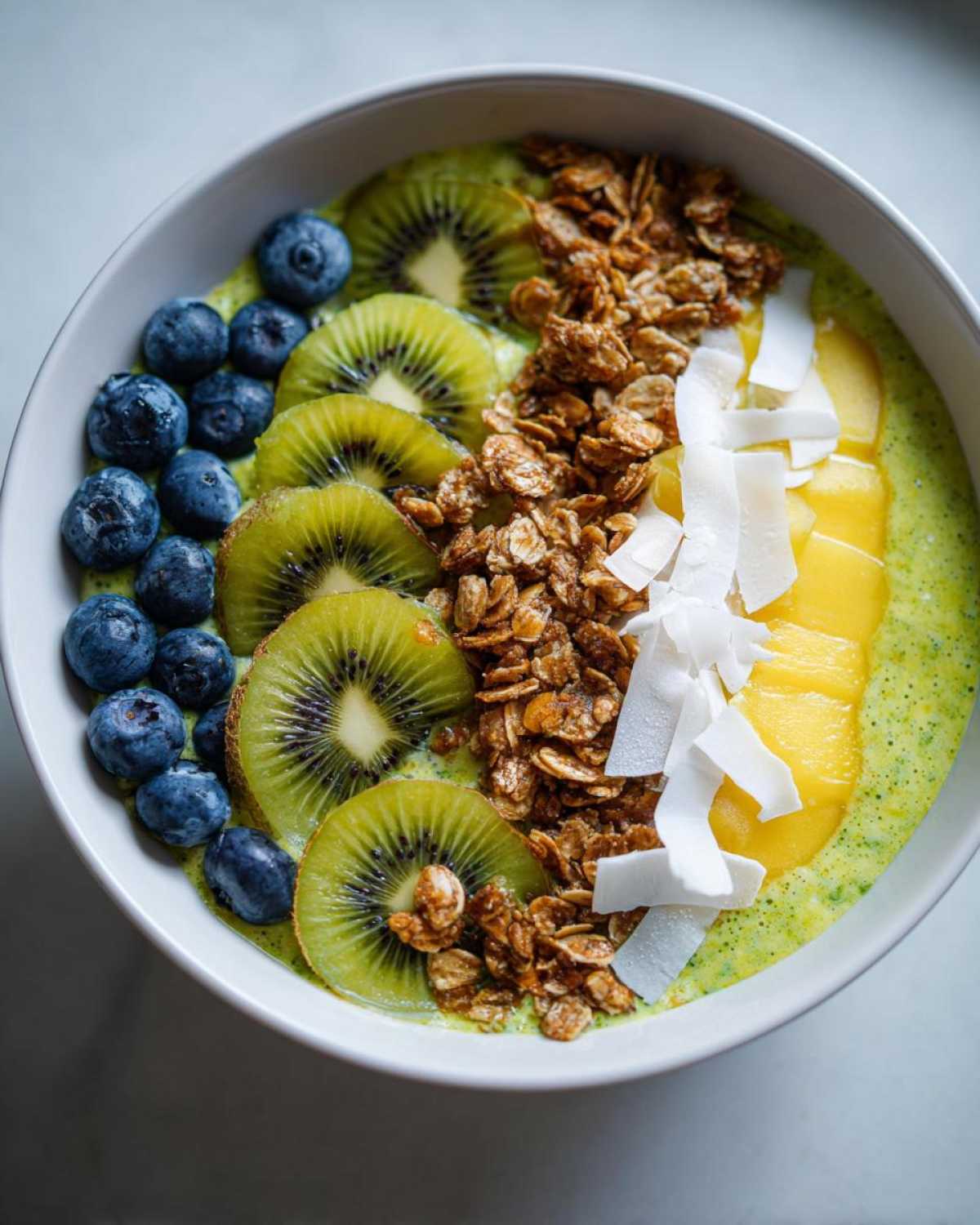 Zbliżenie na zielony Smoothie bowl z owocami: kiwi, borówkami, mango, granolą i wiórkami kokosowymi.