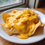 Zbliżenie na nachos polane obficie gęstym, żółtym sosem serowym do nachos na białym talerzu.