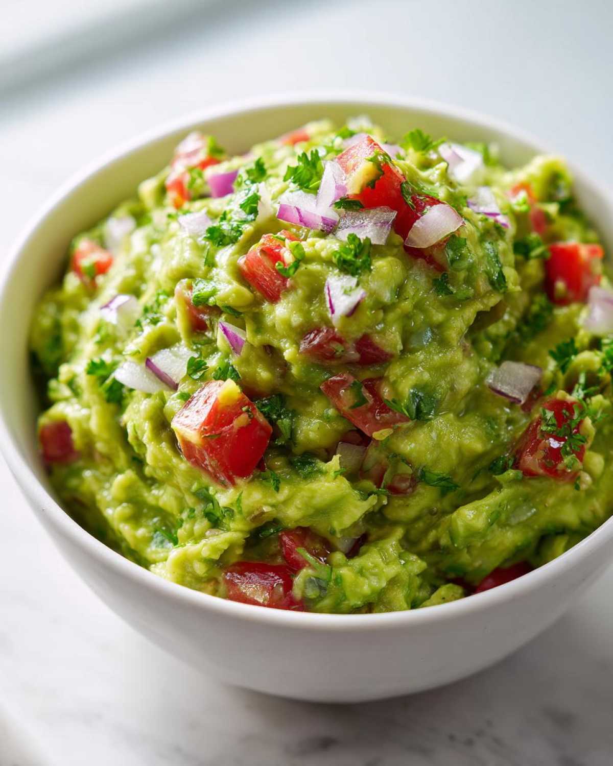 Gęste, zielone Guacamole w białej misce, posypane kawałkami pomidora i czerwonej cebuli.