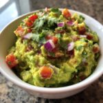 Zbliżenie na miseczkę domowego Guacamole, posypanego kawałkami pomidora, czerwonej cebuli i kolendry.