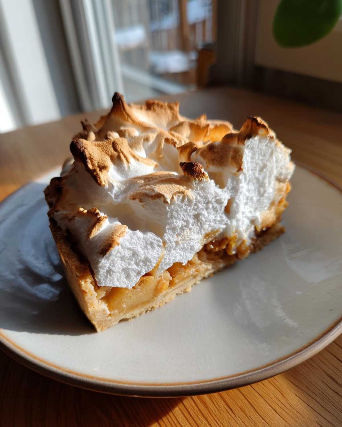 Kawałek puszystej Szarlotki z bezą z przypieczoną górą, podany na jasnym talerzyku.