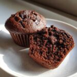 Dwa wilgotne muffiny czekoladowe, jeden przekrojony, z widocznymi kawałkami czekolady na wierzchu i w środku.
