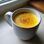 Zbliżenie na kubek wypełniony intensywnie żółtym Złote mleko z kurkumą, posypane cynamonem.