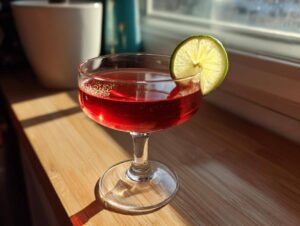 Kieliszek z intensywnie czerwonym drinkiem walentynkowym, udekorowany plasterkiem limonki, stojący na drewnianym parapecie.