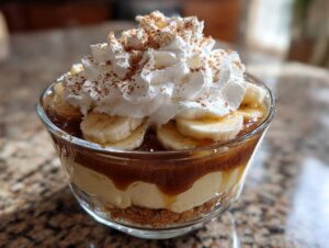 Warstwowy deser bananowy z karmelem, kremem i bitą śmietaną w szklanym pucharku.
