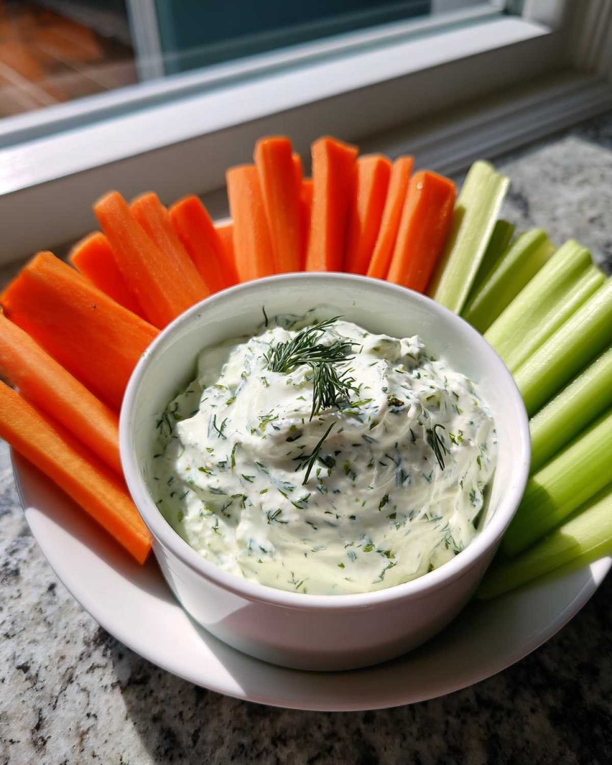 Miseczka kremowego Dip czosnkowy do warzyw otoczona słupkami marchewki i selera naciowego.