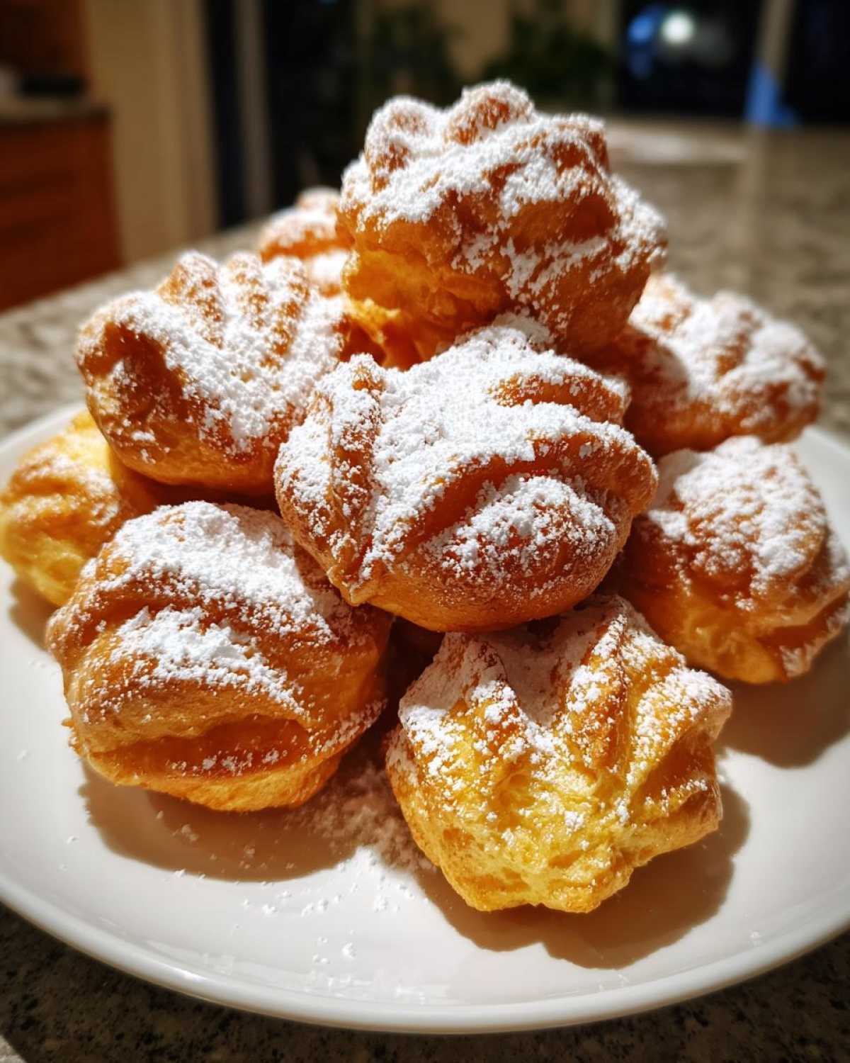 Stos puszystych faworków drożdżowych obficie posypanych cukrem pudrem na białym talerzu.