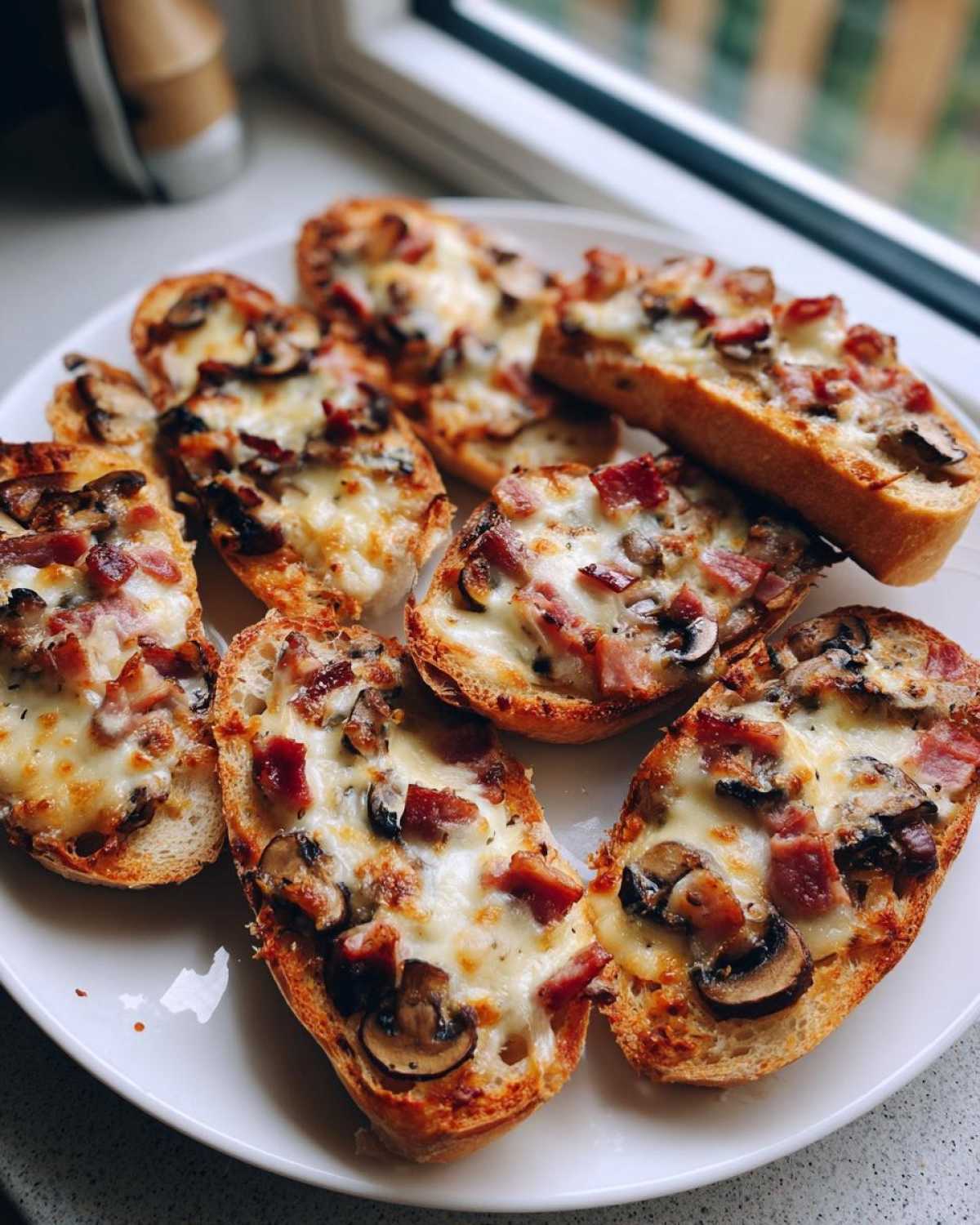 Talerz pełen chrupiących Mini zapiekanki z bagietki posypane serem, boczkiem i pieczarkami.