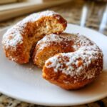 Dwa pączki keto z mąki kokosowej, jeden przełamany, obficie posypane cukrem pudrem na białym talerzu.