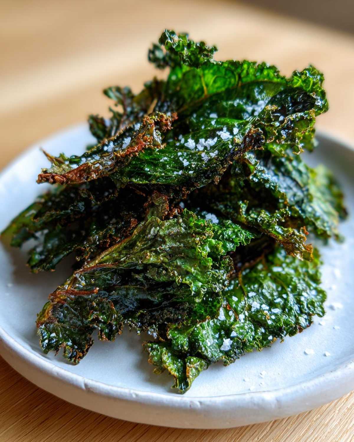 Stos chrupiących, zielonych domowe chipsy z jarmużu posypane kryształkami soli na białym talerzyku.