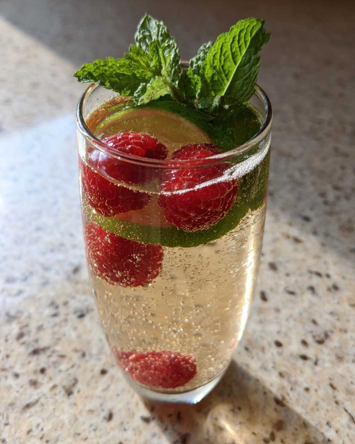 Szklanka orzeźwiającego drinka z Prosecco, udekorowana malinami, plasterkami limonki i listkami mięty.