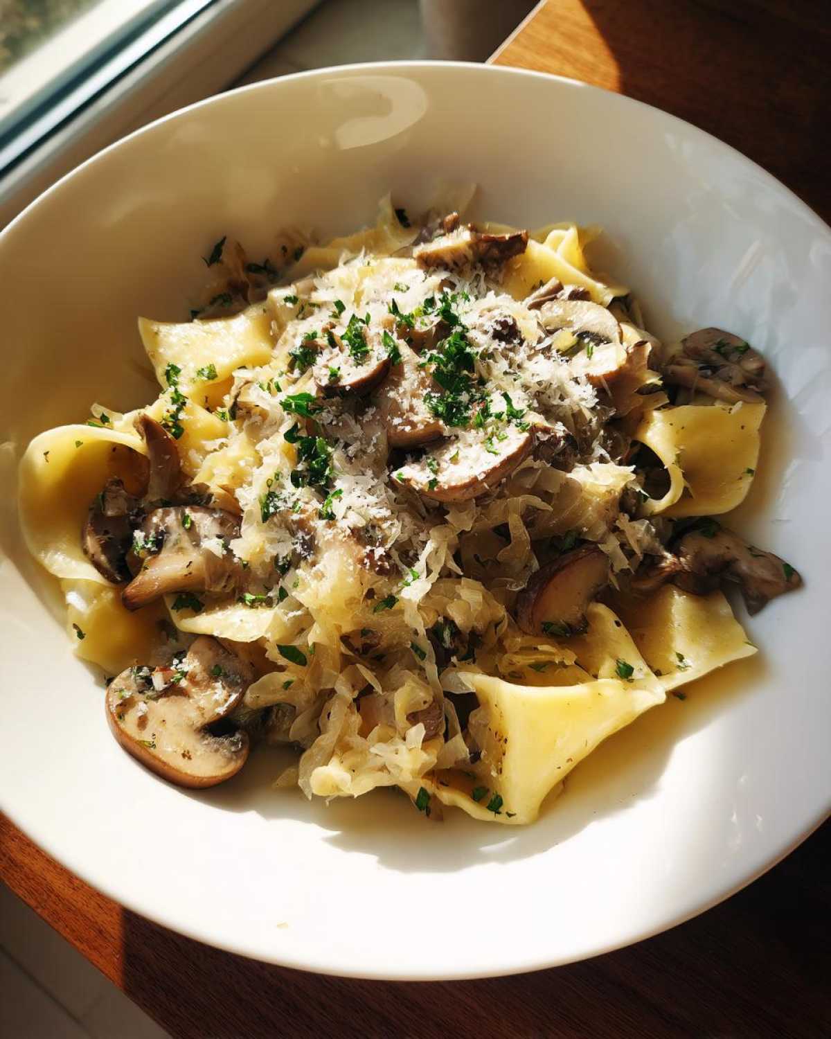 Porcja łazanki z kapustą i pieczarkami posypane serem i pietruszką w białej misce.