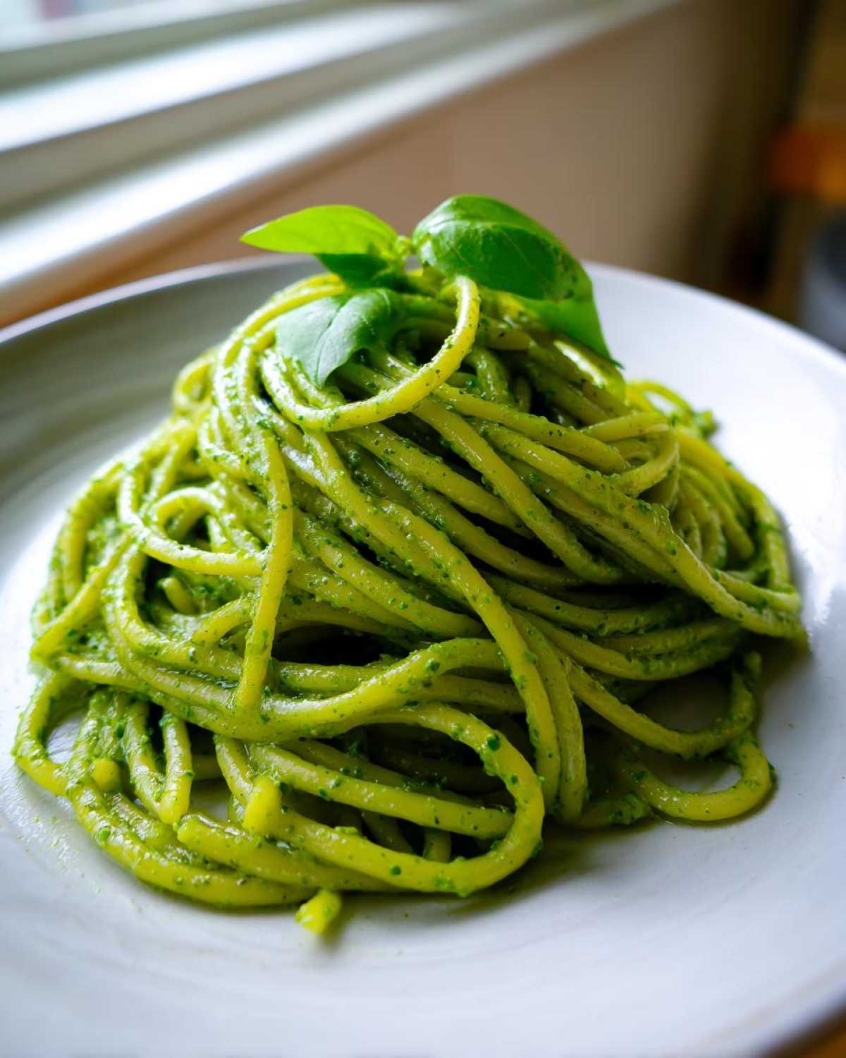 Zbliżenie na porcję makaronu z pesto (zielonym) polanego sosem i udekorowanego świeżymi listkami bazylii.