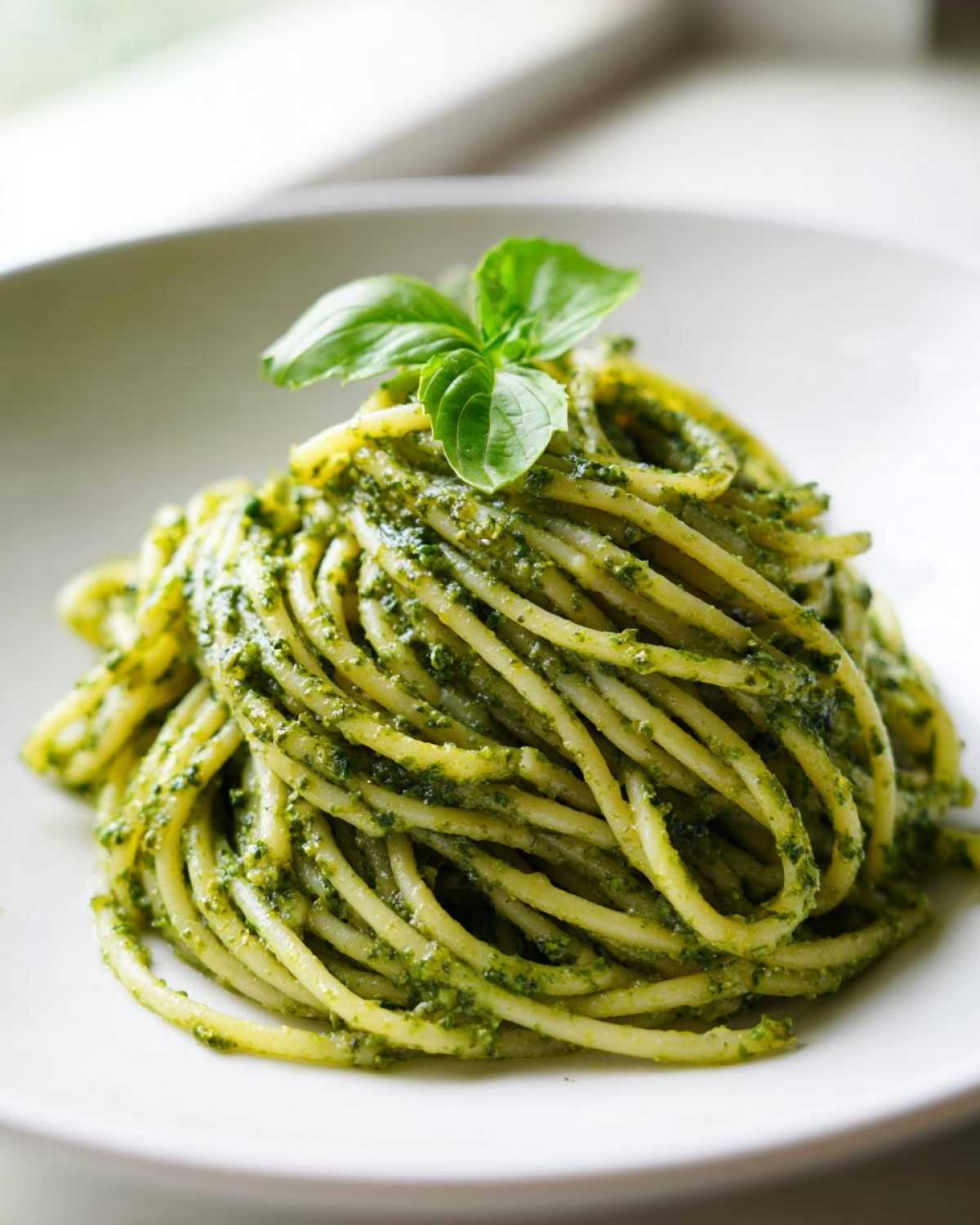 Zbliżenie na porcję makaronu spaghetti obficie polanego zielonym pesto, udekorowanego listkiem bazylii.