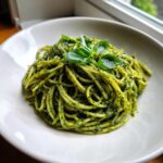 Porcja makaronu spaghetti obficie polana zielonym pesto, udekorowana świeżymi liśćmi bazylii.