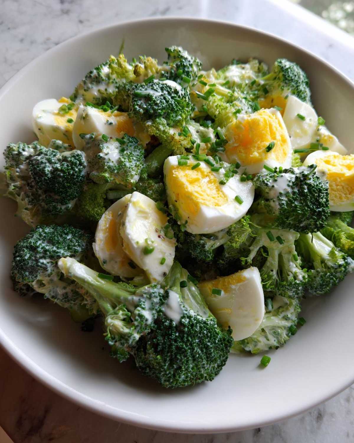 Zbliżenie na miskę pełną sałatki z brokułem, jajkiem i kremowym sosem czosnkowym, posypaną szczypiorkiem.