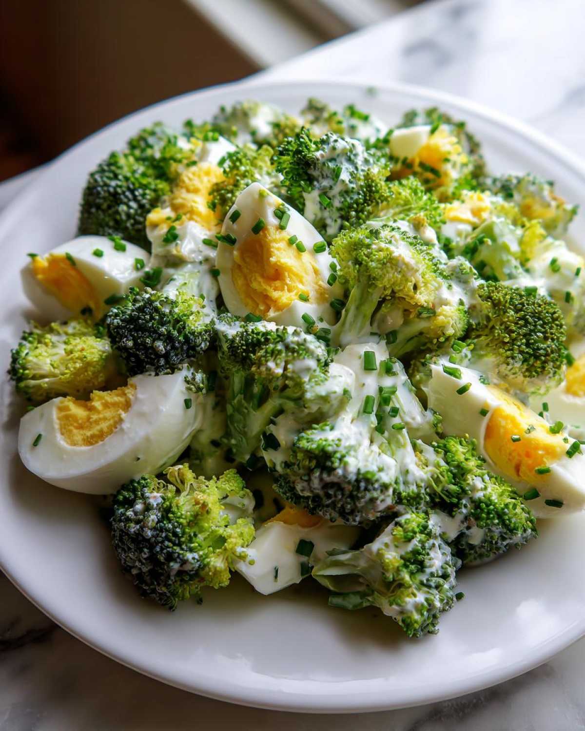 Zbliżenie na talerz pełen sałatki z brokułem, jajkiem i kremowym sosem czosnkowym, posypanej szczypiorkiem.