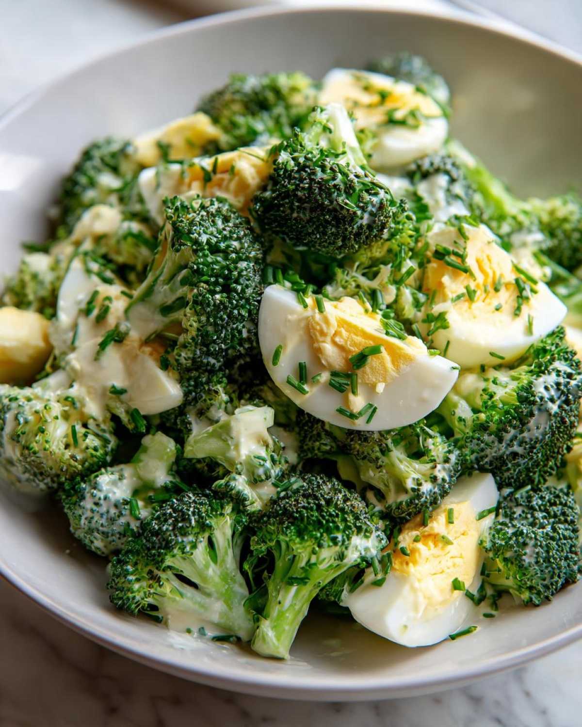 Zbliżenie na miskę pełną sałatki z brokułem, jajkiem i kremowym sosem czosnkowym, posypanej szczypiorkiem.