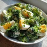 Zbliżenie na miskę Sałatka z brokułem, jajkiem i sosem czosnkowym, polana kremowym sosem i posypana szczypiorkiem.