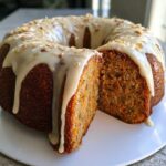 Przekrojona babka marchewkowa polana białym lukrem i posypana orzechami na białym talerzu.