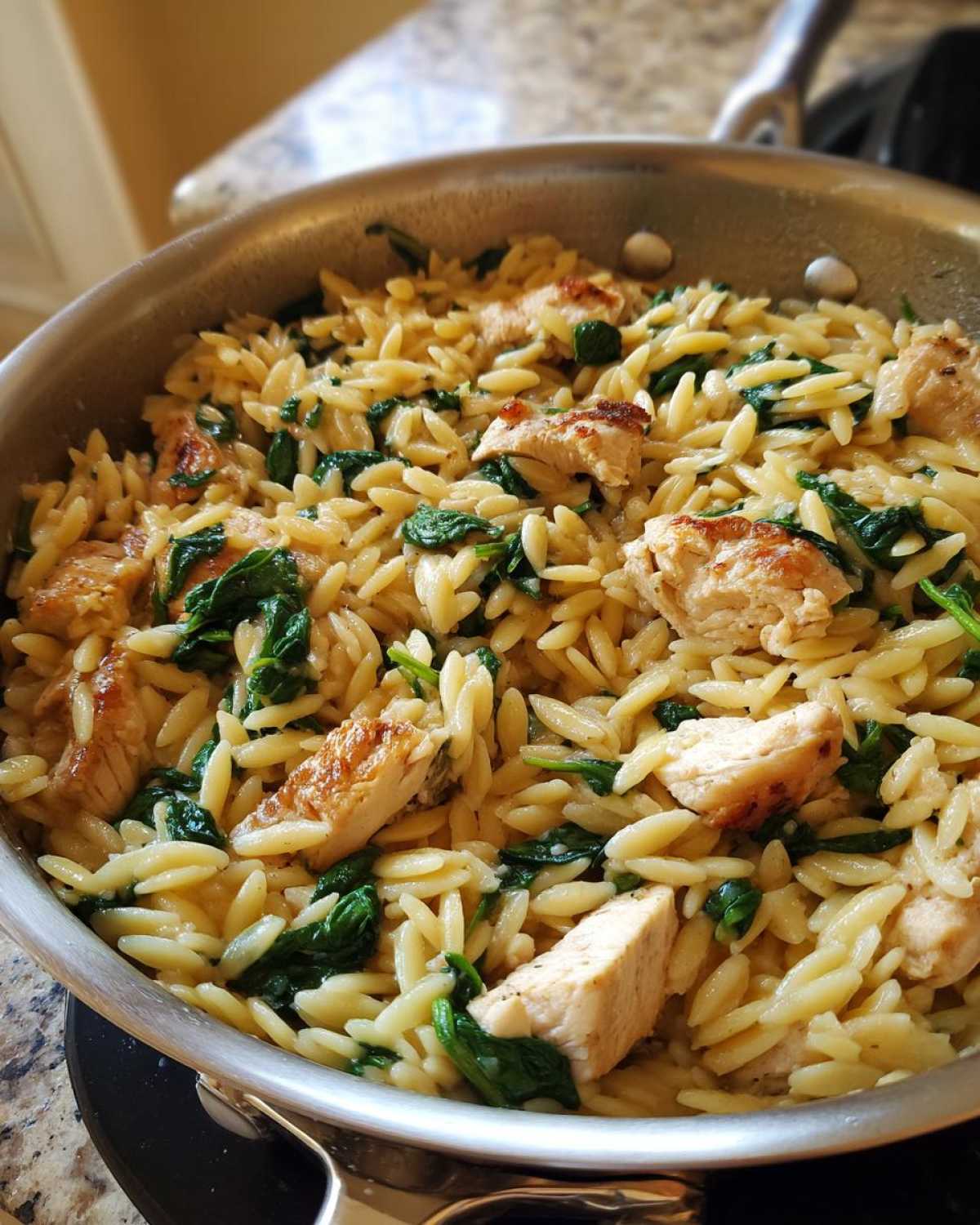 Zbliżenie na kremowy makaron orzo ze szpinakiem i kawałkami kurczaka w metalowej patelni.