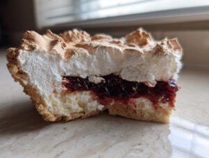 Zbliżenie na przekrojony kawałek Mazurka z bezą i powidłami, ukazujące warstwy ciasta, dżemu i bezy.