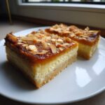 Dwa kawałki Mazurka kajmakowego z kremowym środkiem i migdałami na wierzchu, na białym talerzu.
