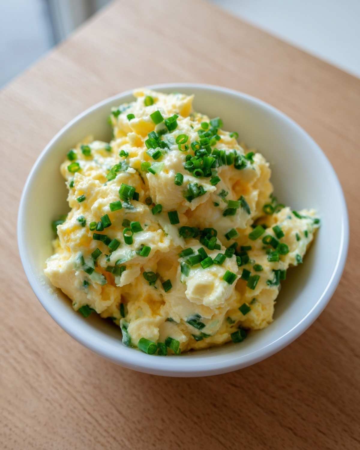 Kremowa pasta z jajek ze szczypiorkiem podana w białej miseczce na drewnianym stole.