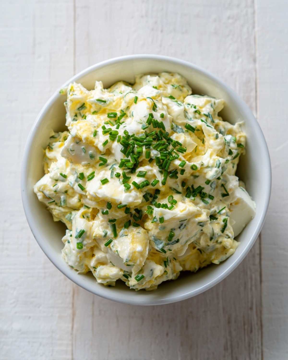 Kremowa pasta z jajek ze szczypiorkiem, podana w białej misce na białym drewnianym tle.