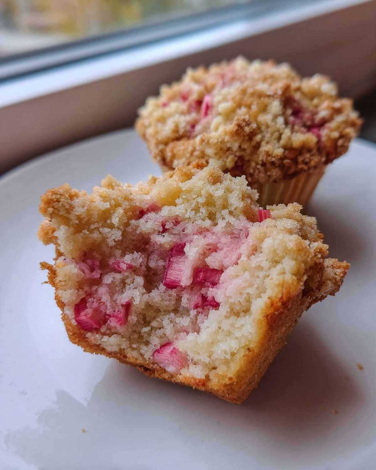 Przekrojone na pół puszyste muffinki z rabarbarem i chrupiącą kruszonką na białym talerzyku.