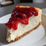 Kawałek kremowego sernika z grubą warstwą błyszczącej polewy malinowej na białym talerzu.