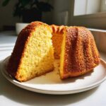 Przekrojona, złocista babka bezglutenowa na białym talerzu, oświetlona słońcem.