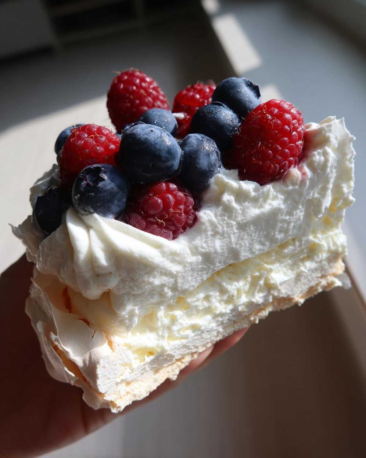 Kawałek Bezy Pavlova z owocami leśnymi, przełożony bitą śmietaną i udekorowany malinami i borówkami.