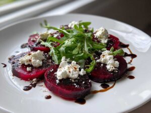 Pięć plastrów gotowanego buraka ułożonych na talerzu, udekorowanych kozim serem, rukolą i sosem balsamicznym. Idealne Carpaccio z buraka z kozim serem.