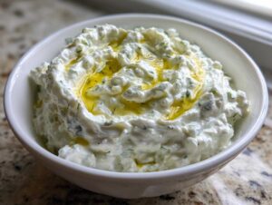 Gęsty sos tzatziki grecki w białej miseczce, polany oliwą z oliwek i udekorowany koperkiem.