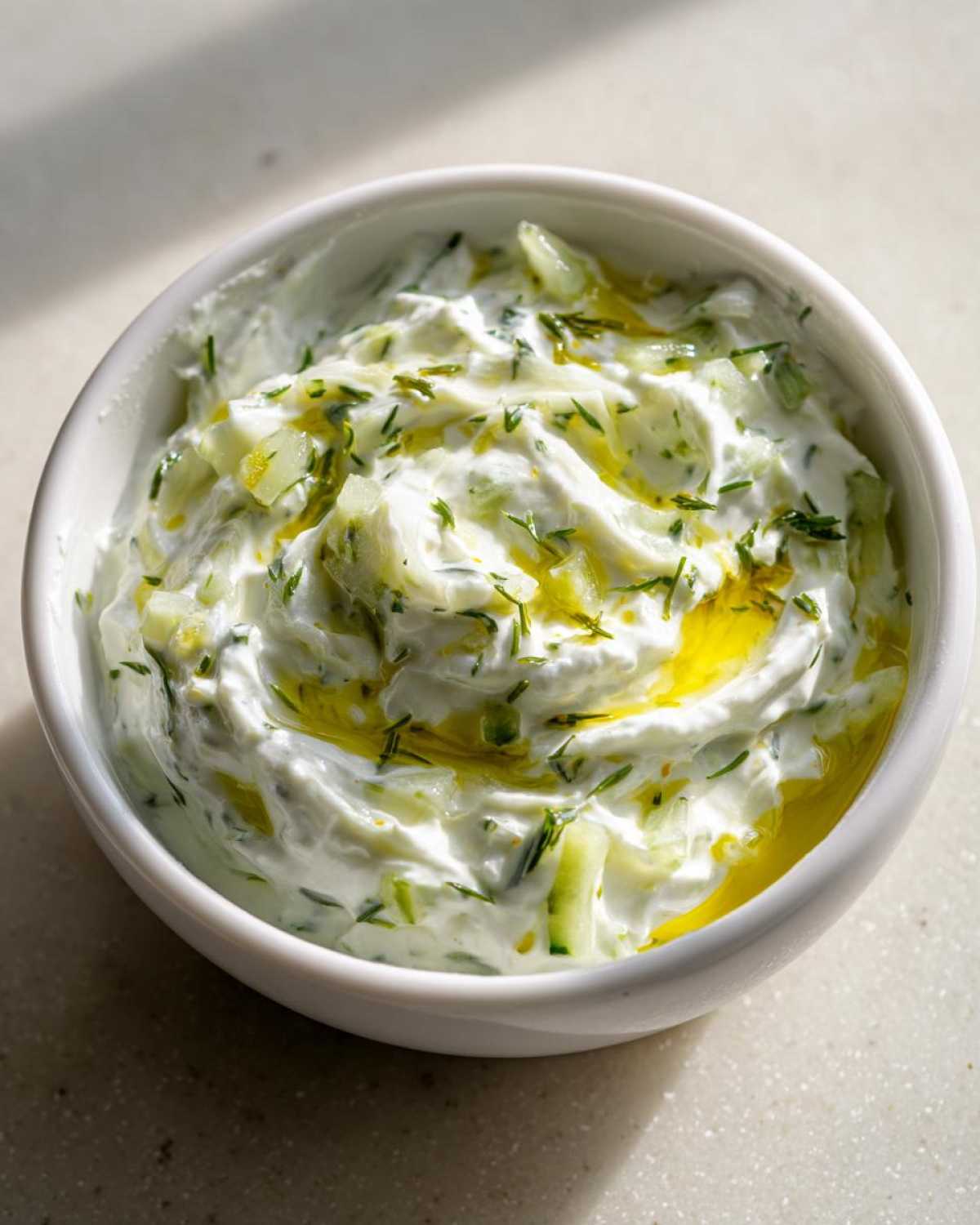 Gęsty sos tzatziki grecki z kawałkami ogórka i oliwą z oliwek w białej misce.
