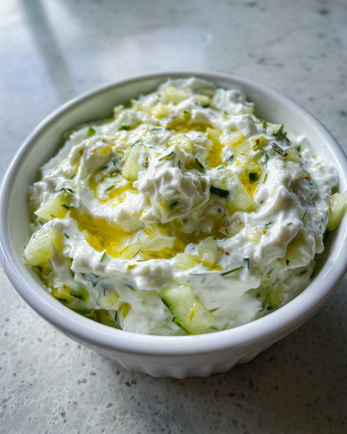 Zbliżenie na gęsty sos tzatziki grecki z kawałkami ogórka i oliwą z oliwek w białej miseczce.