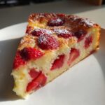 Kawałek puszystego Clafoutis z truskawkami posypany cukrem pudrem, oświetlony słońcem.