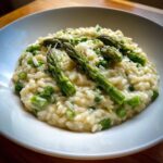 Kremowe Risotto z zielonymi szparagami i groszkiem posypane parmezanem w białej misce.