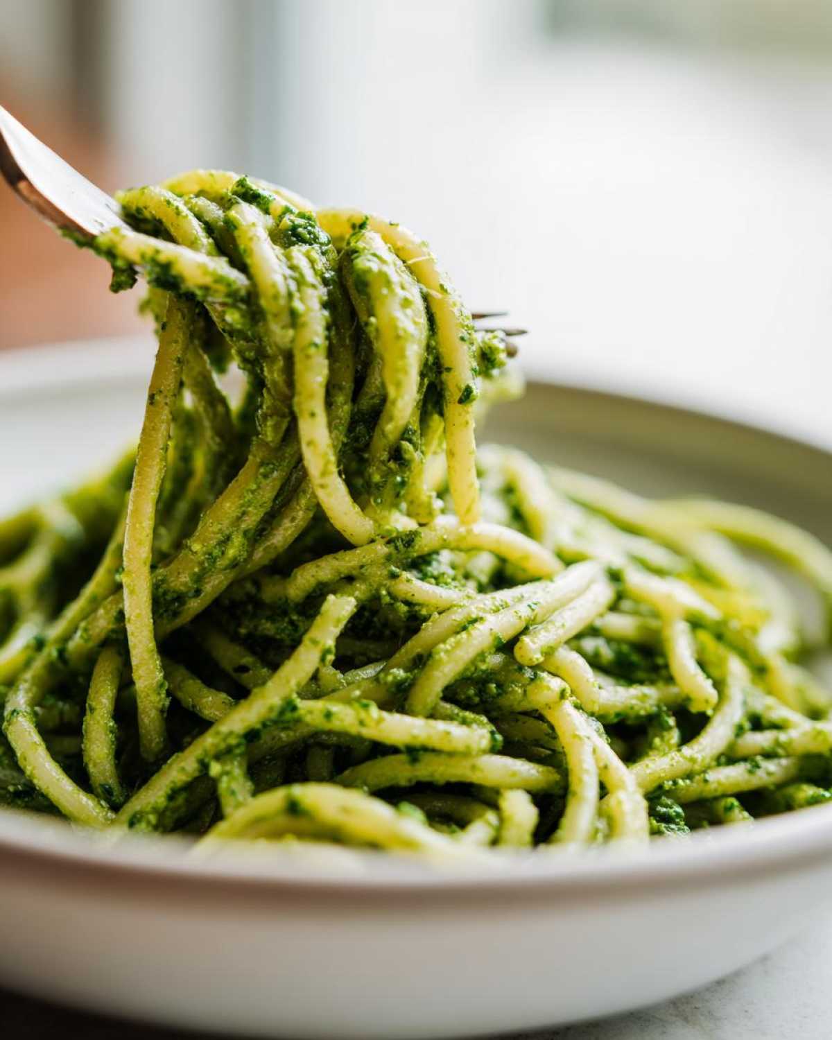 Zbliżenie na makaron typu bucatini polany intensywnie zielonym Pesto z liści botwinki, podnoszony widelcem.