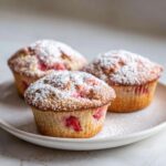 Trzy puszyste muffinki z rabarbarem posypane obficie cukrem pudrem na jasnym talerzu.