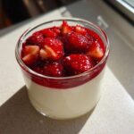 Kremowa panna cotta z truskawkami i sosem truskawkowym w małej szklance.