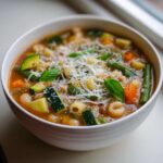 Zbliżenie na miskę pełną Wiosenna zupa minestrone z makaronem, cukinią, fasolką i startym serem.